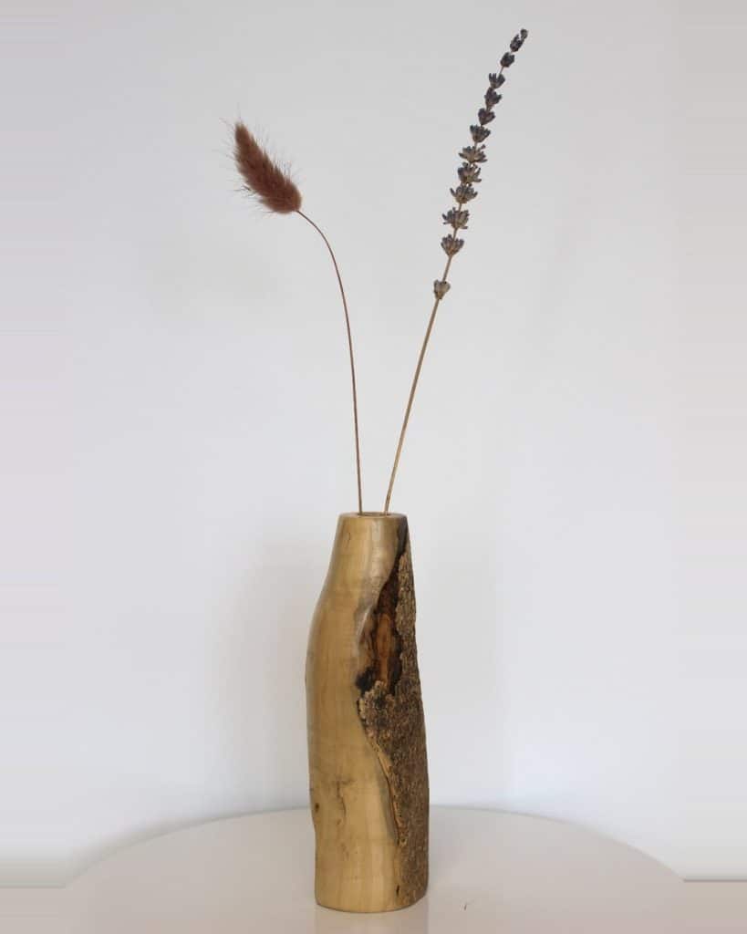 Light wooden vase with bark on one side holding two dried stems: a brown foxtail and a tall lavender spike, on a white surface