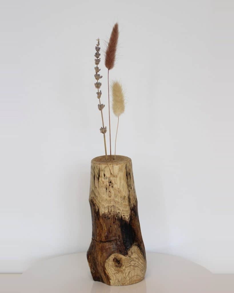 Three dried grass stems in a natural wooden vase on a white surface.