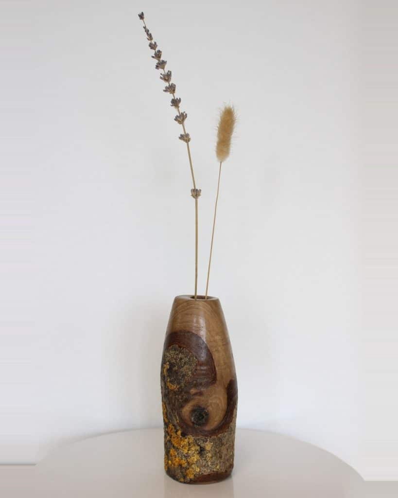 Minimalist still-life: a carved wooden vase holding two dried grasses, one tall lavender-like stem and a fluffy beige inflorescence.